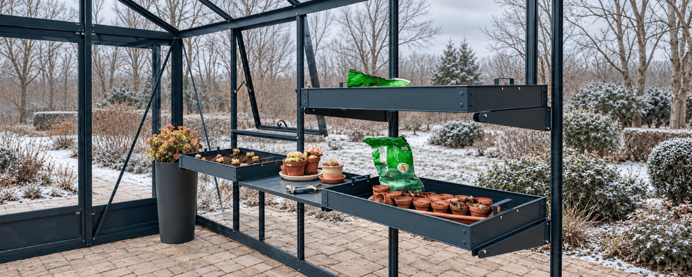 Intérieur d’une serre LAMS avec bacs de jardinage, pots et outils, donnant sur un jardin enneigé.